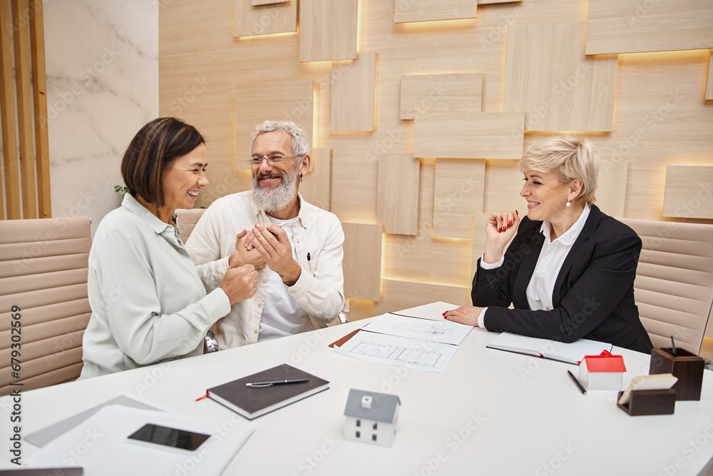 middle aged realtor looking at happy married couple holding key from new house in real estate office middle aged realtor looking at happy married couple holding key from new house in real estate office