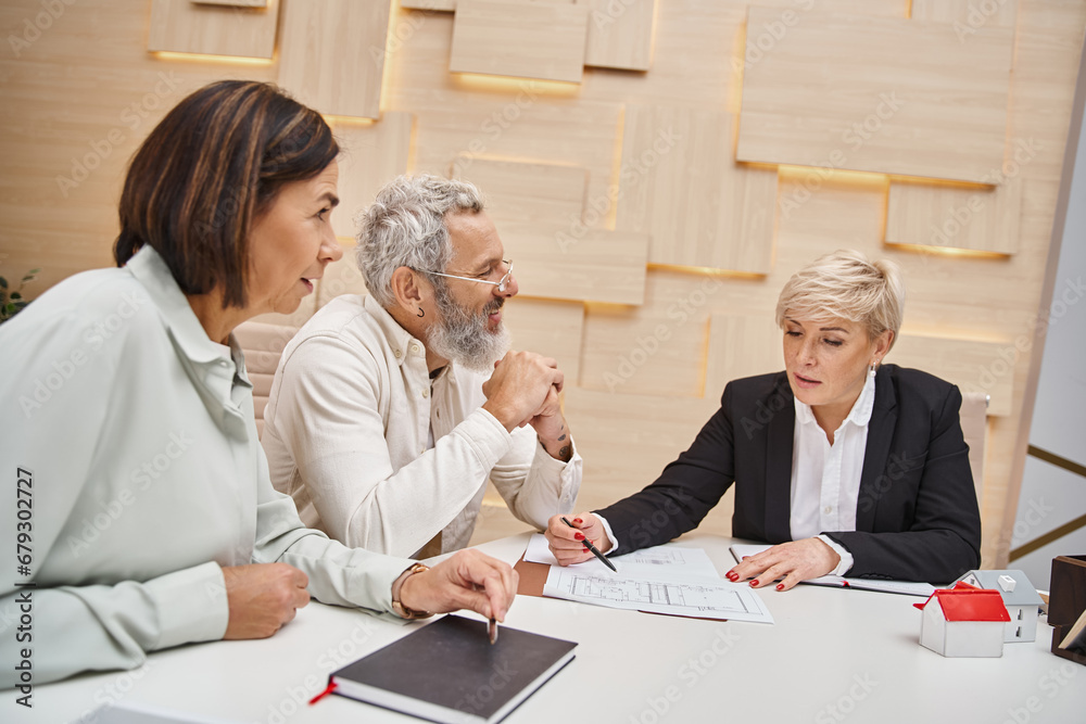 happy middle aged married couple smiling near female realtor and discussing project of new house