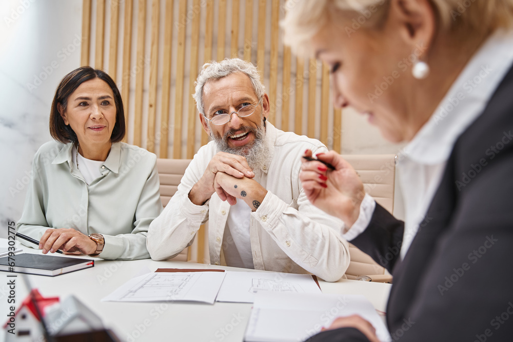 happy middle aged married couple smiling near female realtor and sitting near blueprint of new home happy middle aged married couple smiling near female realtor and sitting near blueprint of new home