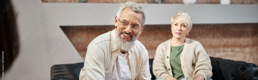 happy bearded man sitting on couch and looking at family consultant near wife during session, banner happy bearded man sitting on couch and looking at family consultant near wife during session, banner