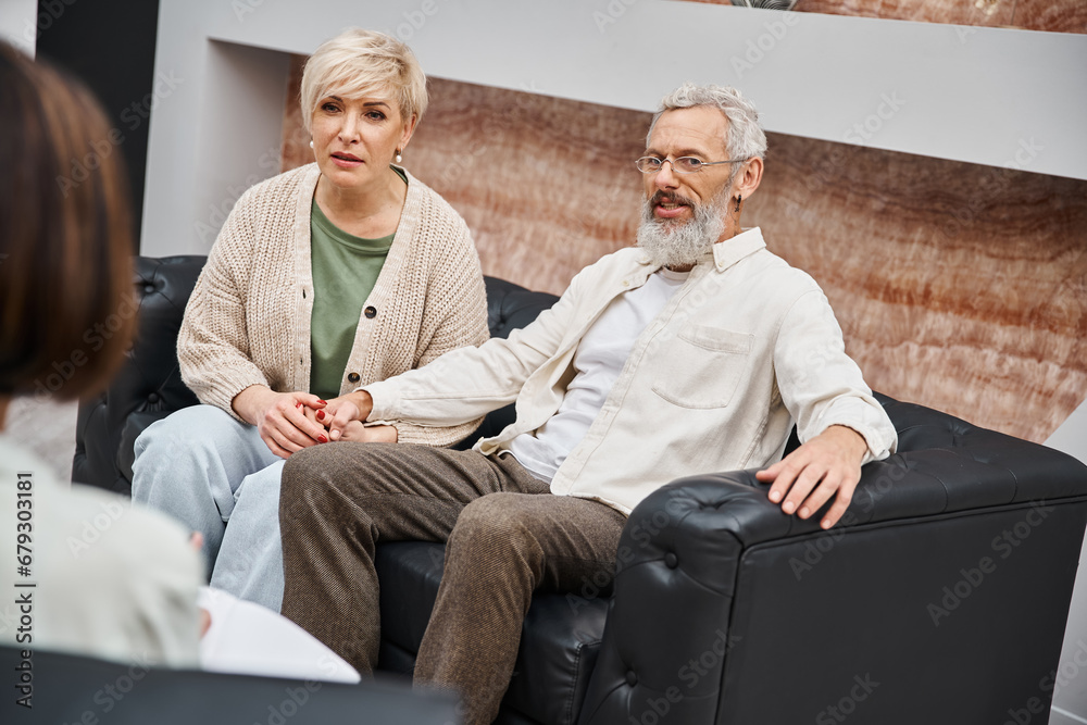 middle aged couple sitting on leather couch and looking at psychologist during family consult