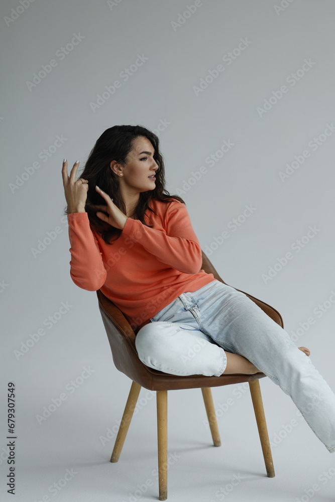 Beautiful woman of 30 years old sitting on chair, white background. Portrait of brunette girl smile. Orange sweater and light jeans. Woman barefeet, thoughtful.