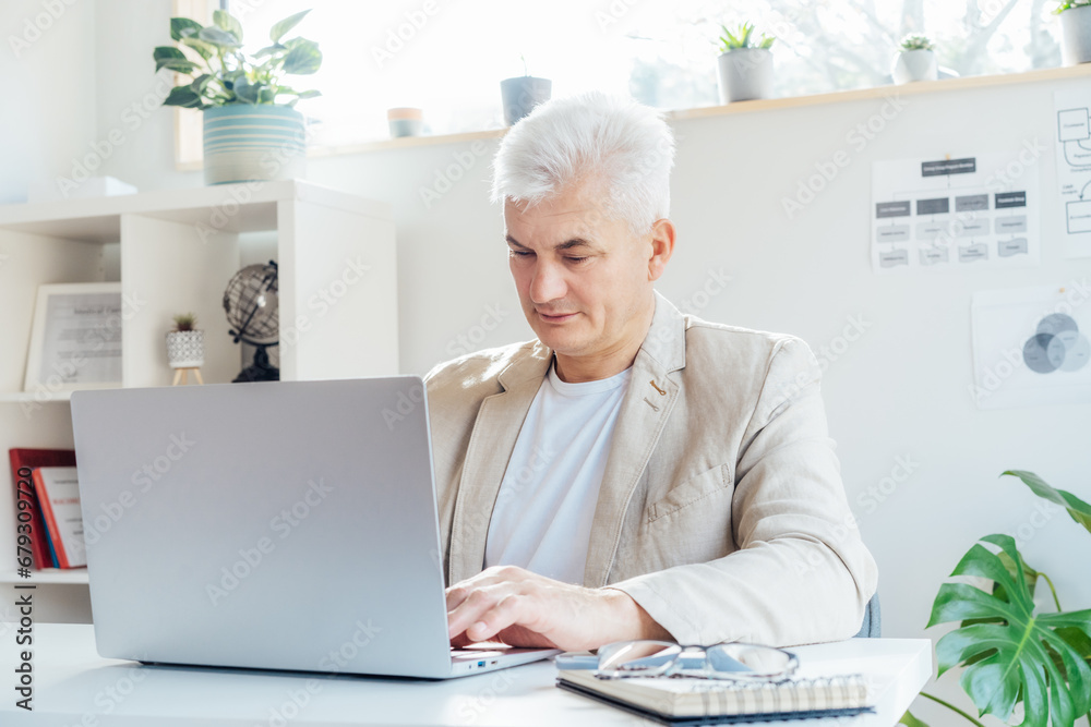Concentrated grey hair middle-aged man working on laptop while sitting at his work place in office. Confident, experienced senior male professional. Corporate manager, entrepreneur manage business. Concentrated grey hair middle-aged man working on laptop while sitting at his work place in office. Confident, experienced senior male professional. Corporate manager, entrepreneur manage business.