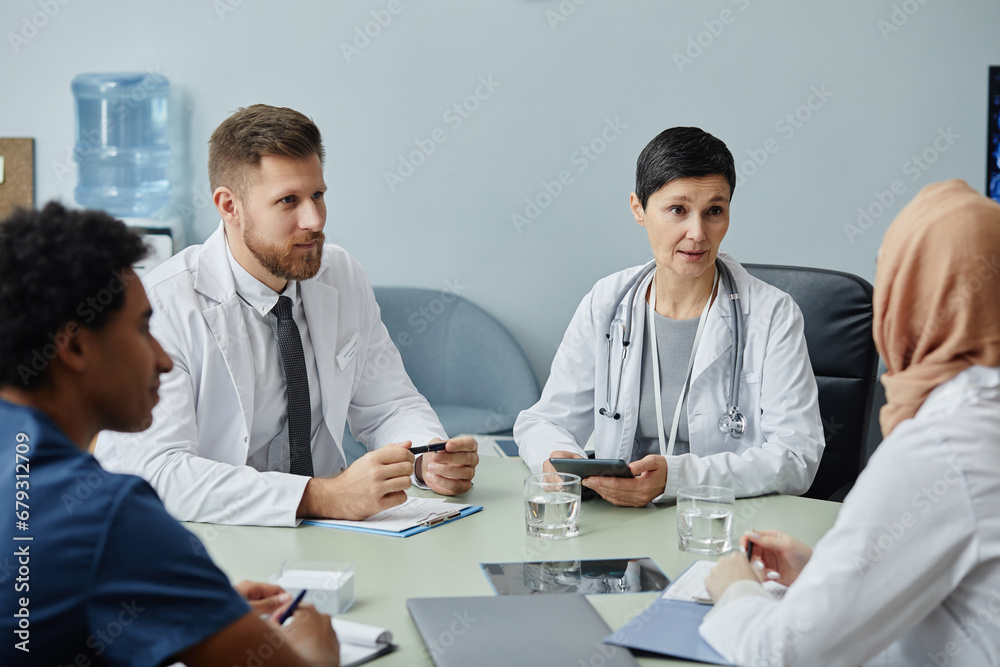 Diverse group of doctors at meeting table in clinic with mature woman speaking to colleagues Diverse group of doctors at meeting table in clinic with mature woman speaking to colleagues