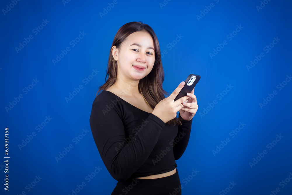 A young Asian woman is happy and smiling while using a smartphone on a blue background A young Asian woman is happy and smiling while using a smartphone on a blue background