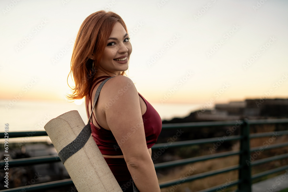 Cheerful plus-size woman carrying a yoga mat, ready for a sunset yoga session by the sea, embodying a body-positive spirit.