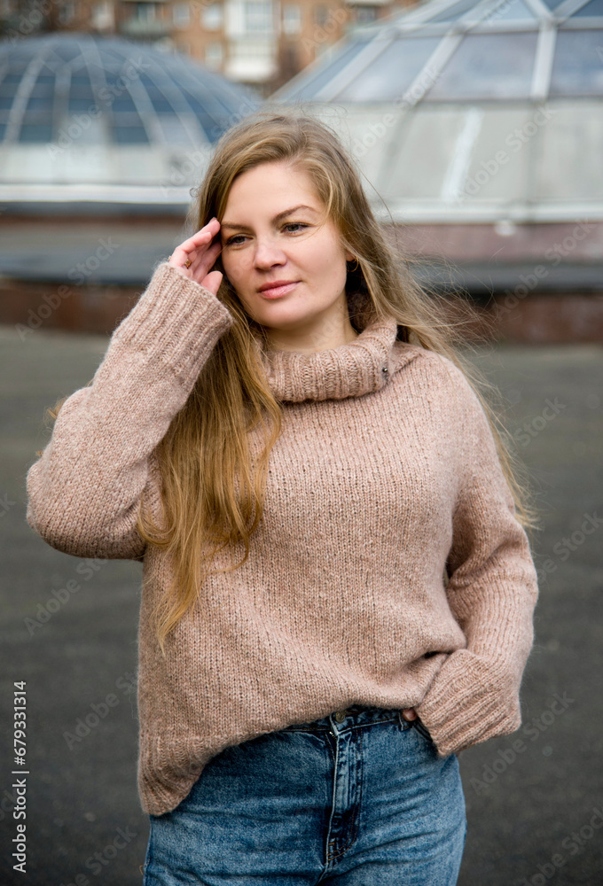 portrait of beautiful blonde smiling woman with long hair in beige handmade knitted sweater and blue jeans posing on street on autumn day portrait of beautiful blonde smiling woman with long hair in beige handmade knitted sweater and blue jeans posing on street on autumn day