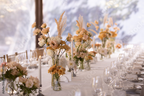 A large, long, decorated, wooden table and chairs, covered with a white tablecloth with dishes, flowers, candles.
