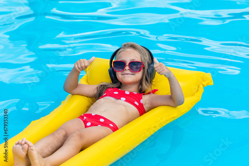 Happy little girl listening to music at pool. Closeup portrait of Caucasian little girl in swimsuit, listening to music in wireless headphones, smiling cute in pool. Tropical summer vacation concept