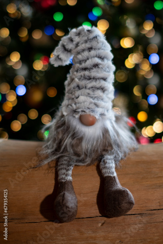 Petit Père-Noël gris et blanc, avec un bonnet gris et blanc très grand qui cache ses yeux, petit nez avec une barbe blanche, assis sur un tronc d'arbre devant des décorations lumineuses  de Noël 