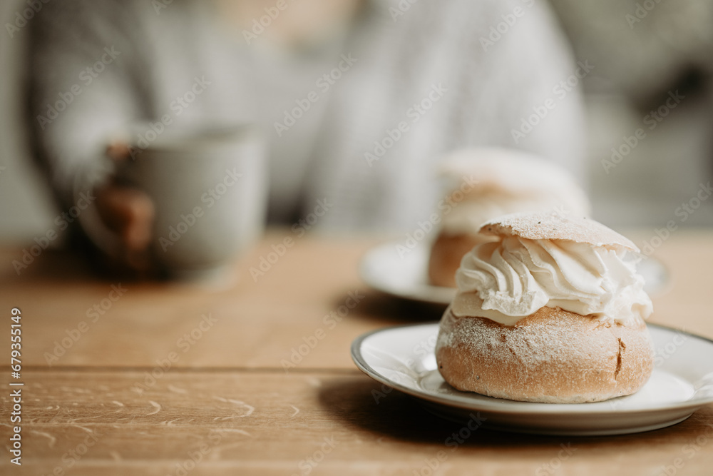Traditional Swedish pastry, known as semla, on a plate at wooden table. In the background, out of focus, is a person with grey sweater, holding a coffee cup. Photo taken in Sweden. - obrazy, fototapety, plakaty 