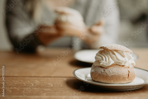 Focus on traditional Swedish pastry, semla, on a plate at wooden table. In the background, out of focus, a person is about to eat pastry. Photo taken in Sweden. photo