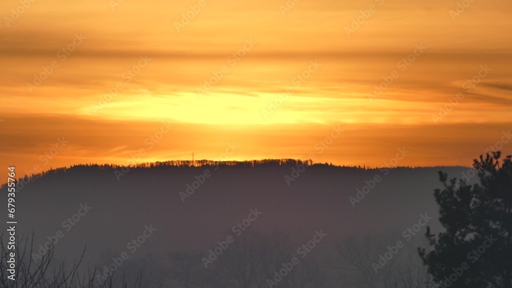 Orange morning sky with clouds and yellow sunlight before sunrise over dark horizon of Silesian Beskid mountains in Poland with forests and Christian cross.