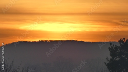 Orange morning sky with clouds and yellow sunlight before sunrise over dark horizon of Silesian Beskid mountains in Poland with forests and Christian cross.