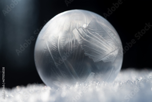 Close-up of a shimmering frozen soap bubble lying on snow crystals. The background is dark. The light shines from the side.