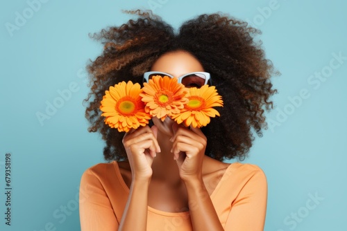 Playful positive young African American woman in funny glasses covering her face with orange gerberas, enjoying spring, fresh flowers, having fun. Isolated on blue background.