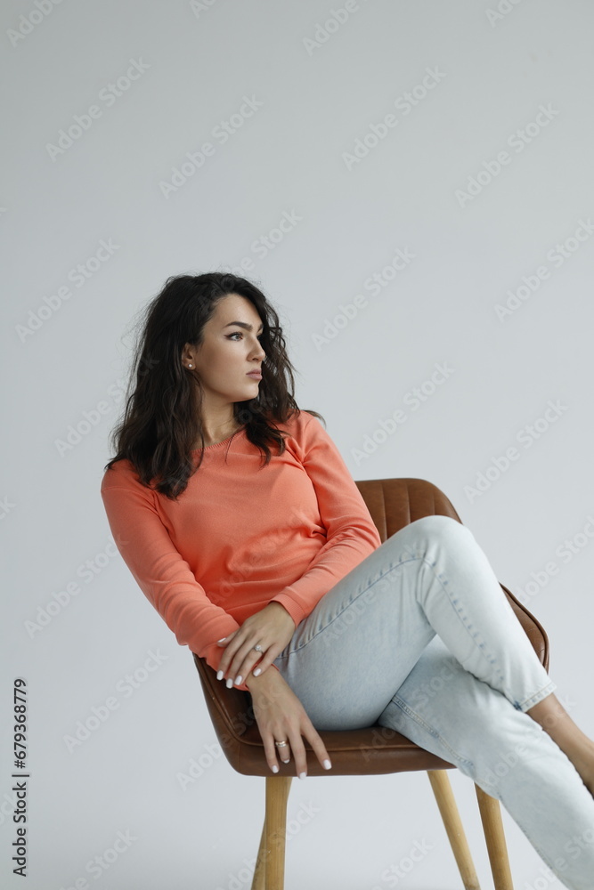 Beautiful woman of 30 years old sitting on chair, white background. Portrait of brunette girl smile. Orange sweater and light jeans. Woman barefeet, thoughtful. Beautiful woman of 30 years old sitting on chair, white background. Portrait of brunette girl smile. Orange sweater and light jeans. Woman barefeet, thoughtful.