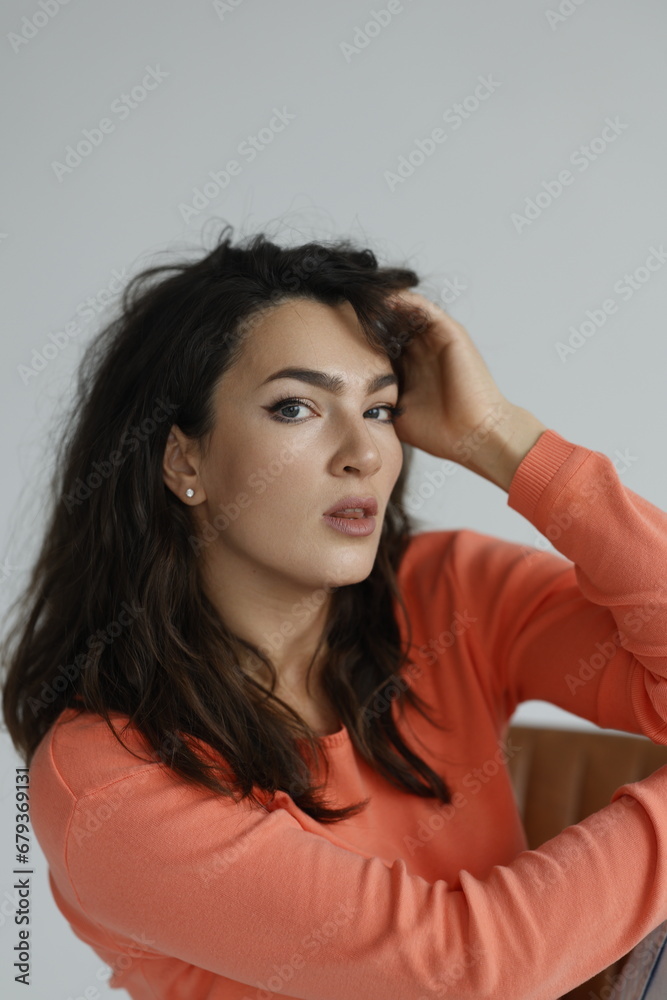 Beautiful woman of 30 years old sitting on chair, white background. Portrait of brunette girl smile. Orange sweater and light jeans. Woman barefeet, thoughtful. Beautiful woman of 30 years old sitting on chair, white background. Portrait of brunette girl smile. Orange sweater and light jeans. Woman barefeet, thoughtful.