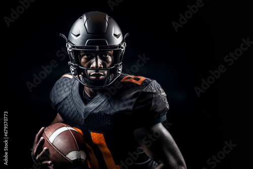 Studio shot of professional American football player in black jersey holding ovoid ball in his hand. Determined, powerful, skilled African American athlete wearing helmet. Isolated in black background
