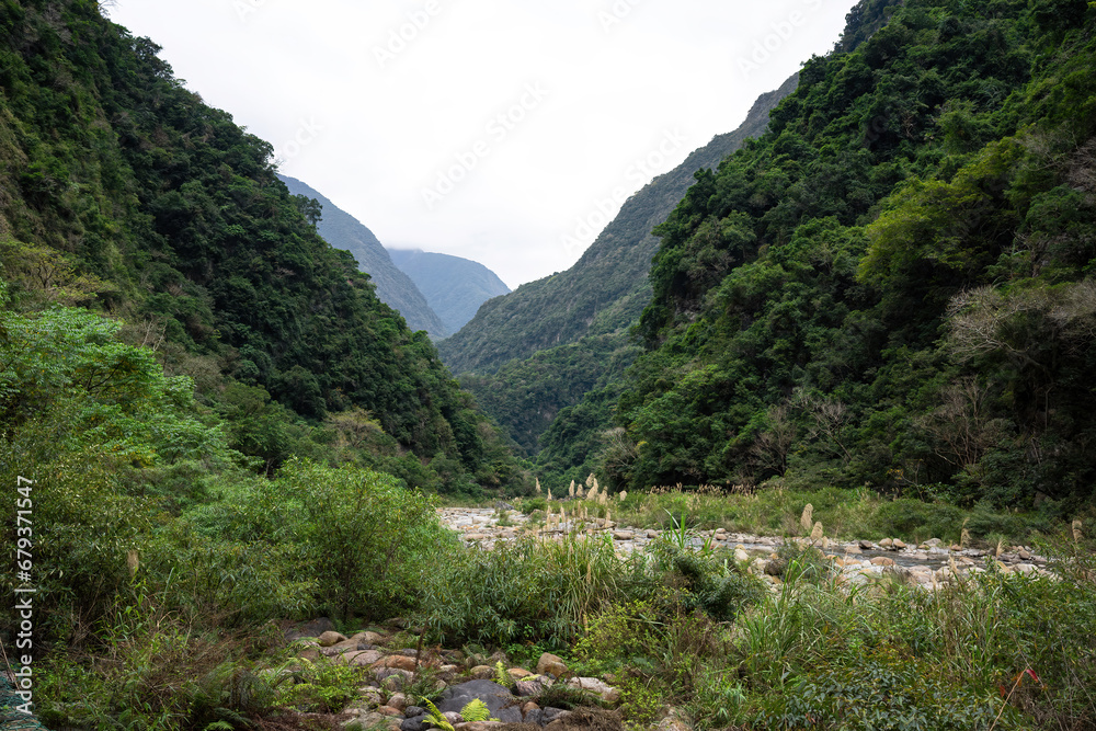 A wide-angle view captures the lush greenery of a valley with a riverbed, nestled between towering green mountains under a soft, cloudy sky.