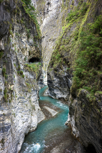 The dramatic landscape of a river gorge with a visible cave entrance is framed by steep, verdant cliffs, offering a glimpse into the rugged terrain's beauty.