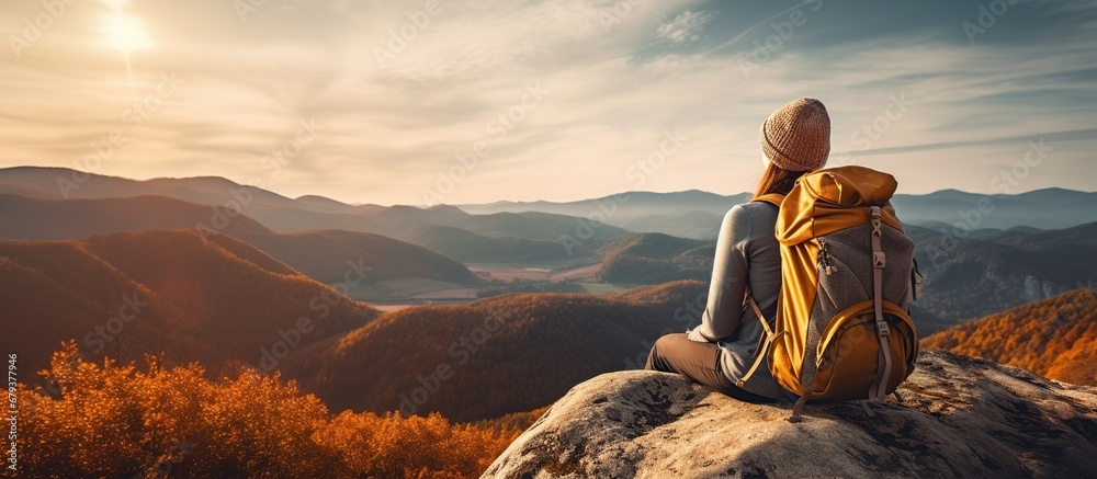 Back view young woman wearing a backpack and sit on stone in mountain at sunset - obrazy, fototapety, plakaty 