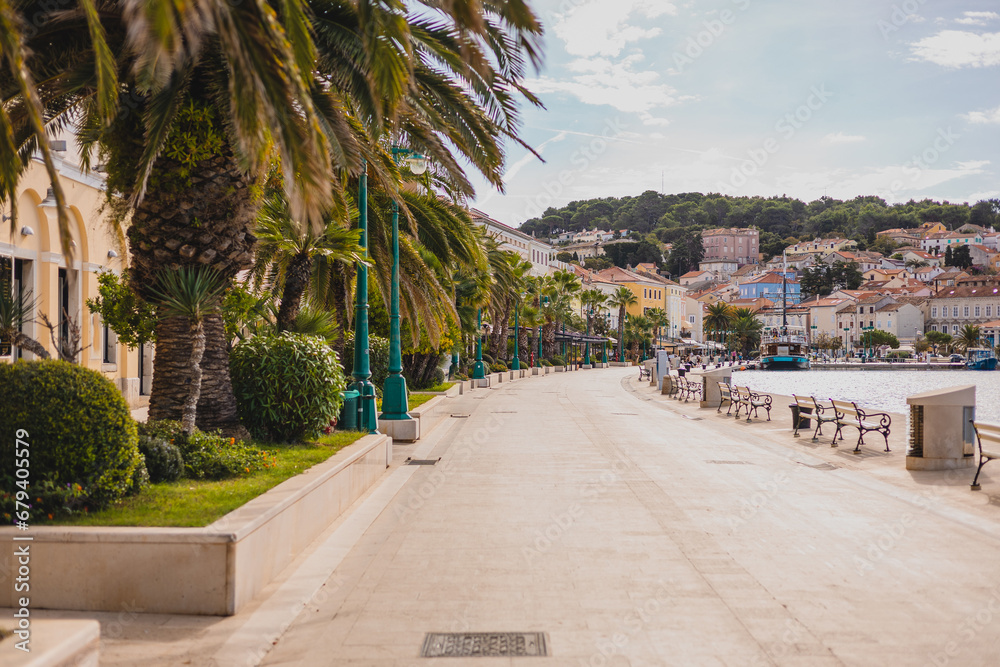 Promenade with palms and other trees and houses in the background in the city of Mali Losinj, on croatian island of Losinj, on a sunny day in autumn. Almost no people visible.