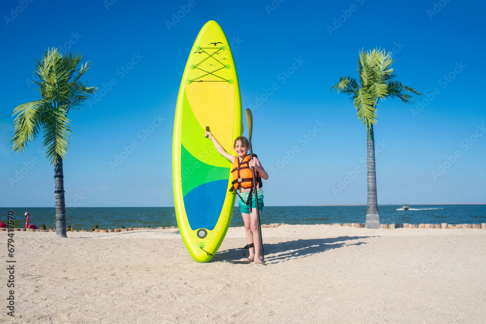 happy 10 year old boy holding a green sup board and paddle for sap boarding on a sunny day at the seaside. a small child in a life jacket is standing with his SUP board on a sandy beach 