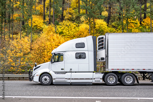 Industrial grade white big rig bonnet semi truck transporting cargo in reefer semi trailer running on the wide highway road with autumn yellow forest on the hill photo
