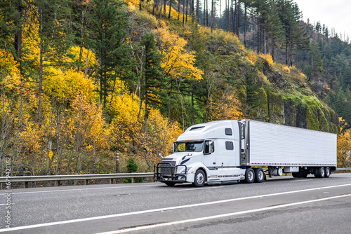 Pro industrial big rig white semi truck with grille guard transporting cargo in refrigerated semi trailer driving on the autumn road with yellow trees on the hill