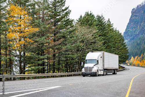 Professional long hauler big rig white semi truck transporting cargo in refrigerator semi trailer driving on wide autumn road along the river in Columbia River Gorge photo