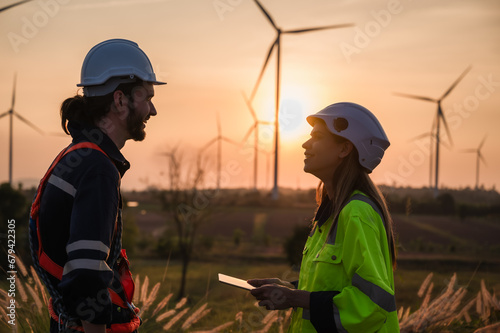 Two professional maintenance engineers in uniform and safety workwear using tablet to checklist repair wind turbines on windmill farm site.