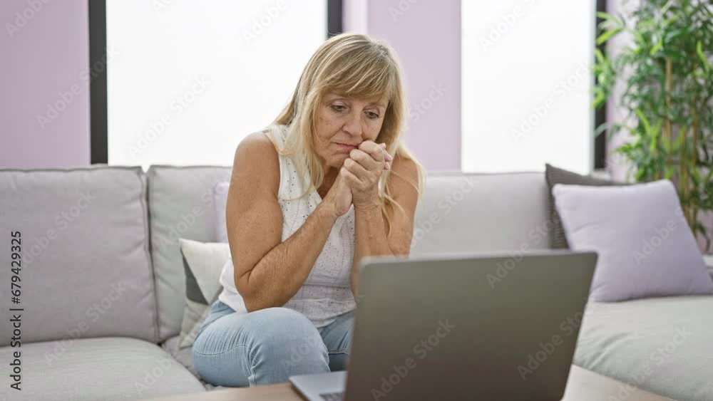 In her cozy home, a serious and focused middle age blonde woman is sitting on the sofa, totally absorbed, using her laptop in the living room's welcoming interior. In her cozy home, a serious and focused middle age blonde woman is sitting on the sofa, totally absorbed, using her laptop in the living room's welcoming interior.