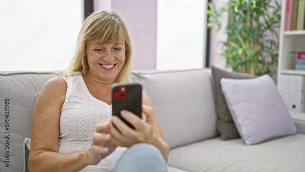 Beautiful middle-aged blonde woman sitting on a sofa at home, enjoying the joy of texting on her smartphone, a cheerful expression of happiness on her face Beautiful middle-aged blonde woman sitting on a sofa at home, enjoying the joy of texting on her smartphone, a cheerful expression of happiness on her face
