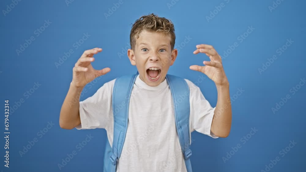 Adorable blond boy student standing, screaming loudly with serious expression over isolated blue background, expressing anger, frustration