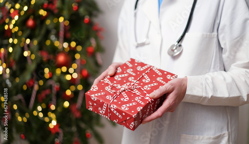 Medical banner concept for Christmas and New Year.Female doctor in white coat holds stethoscope and gift box wrapped in red paper against background of beautifully decorated Christmas tree.Copy space.