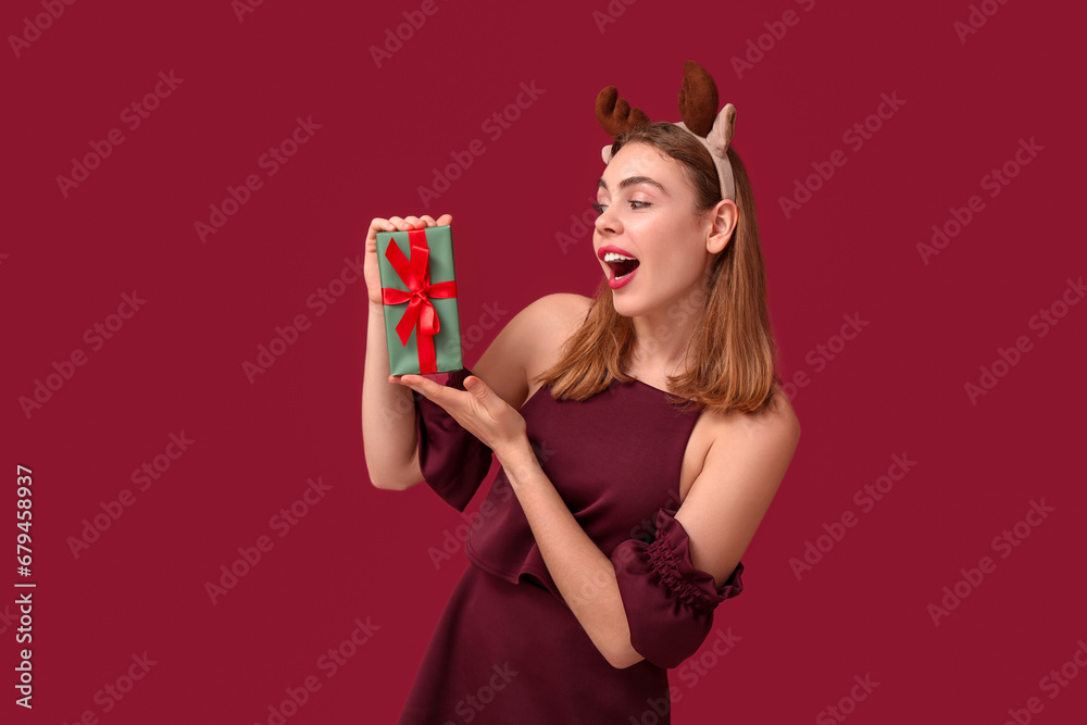Beautiful young woman in reindeer horns with Christmas gift box on red background Beautiful young woman in reindeer horns with Christmas gift box on red background