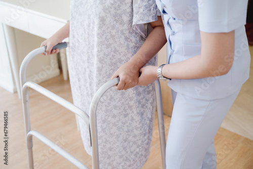 A caring specialist carer observes and supports a mature older female practice walking slowly with a walker at a nursing home.