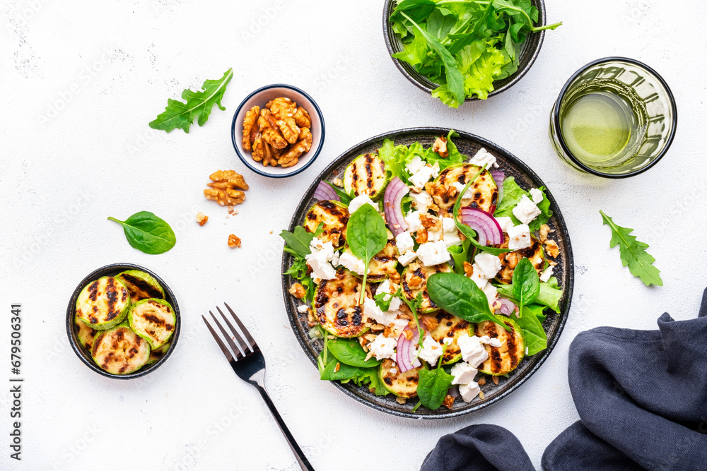 Tasty fresh salad with grilled zucchini, feta cheese, walnuts, red onion and spinach on white table background, top view