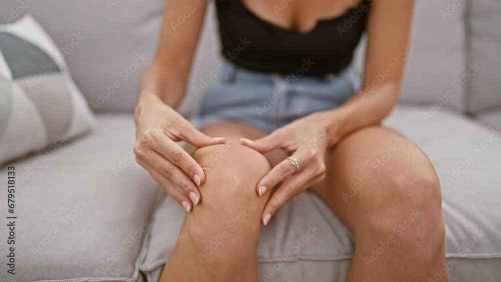 Striking young hispanic woman with beautiful short hair-face etched in pain-touches her injured knee while sitting on sofa in the comfort of her home. Striking young hispanic woman with beautiful short hair-face etched in pain-touches her injured knee while sitting on sofa in the comfort of her home.