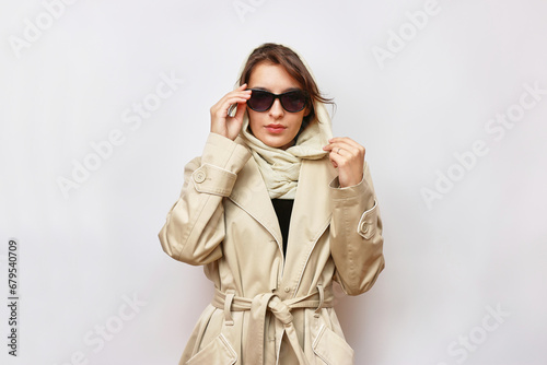 A beautiful girl in sunglasses is dressed in a light autumn raincoat and a scarf.