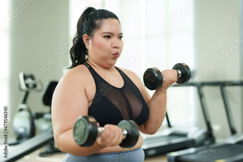 Chubby Asian woman Do a seated exercise by lifting dumbbells with both arms. Trying to lose weight Serious expression Committed to exercising Overweight Asian woman exercising in the gym.