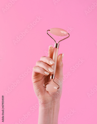 Woman's hand holds a massage jade facial roller on a pink background. Anti-aging therapy