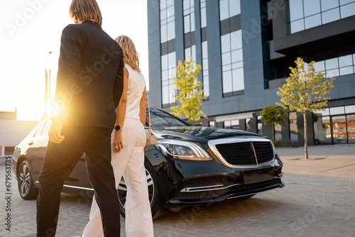 Businessman and businesswoman walk to a luxury black car near hotel or office building on sunset. Concept of transportation and business travel © rh2010