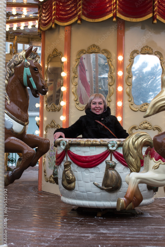 A woman in a winter fur coat rides a carousel in the winter park on the square. New Year's walk around the square