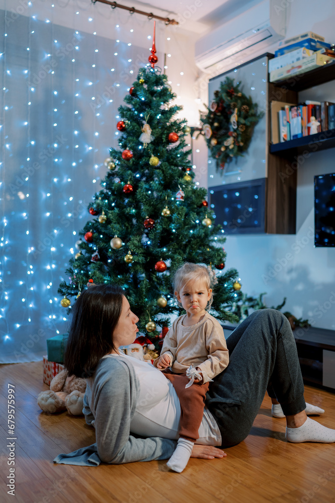 Little girl with pursed lips sits on her mom stomach lying on the floor near a decorated Christmas tree