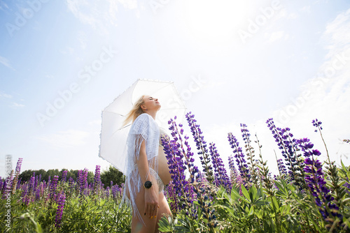 A cute pregnant girl in light underwear and a cape, with light short hair with a white umbrella in her hands, stands and rests in a picturesque meadow with purple blossoms.