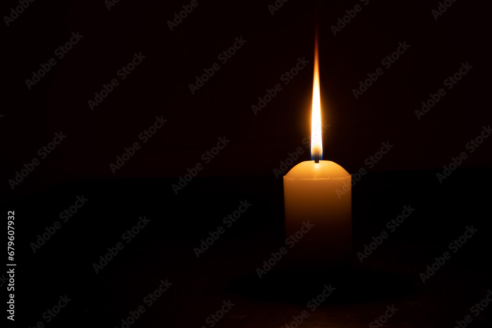A single burning candle flame or light glowing on a yellow candle isolated on red or dark background on table in church for Christmas, funeral or memorial service - obrazy, fototapety, plakaty 