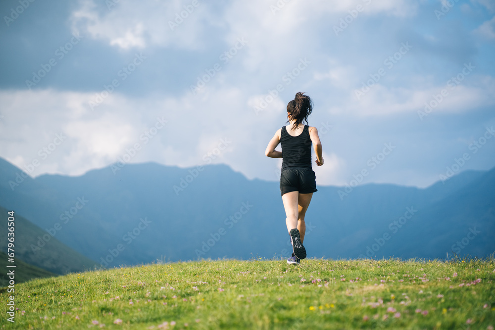 Woman trail running through alpine landscape in morning light, view of mountains behind her Woman trail running through alpine landscape in morning light, view of mountains behind her