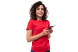 young slender leader woman with black curly shoulder-length hair dressed in a red basic t-shirt holding a smartphone